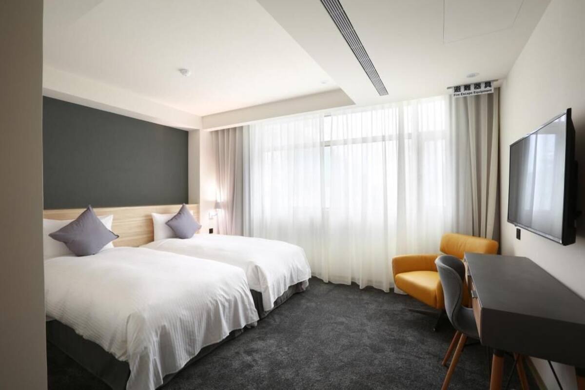 A hotel room with two beds, a desk, a chair, a TV, and a large window with curtains.