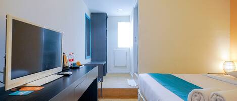 Quarto de hotel com cama, escrivaninha com televisão, luminária de cabeceira e vista para o corredor.