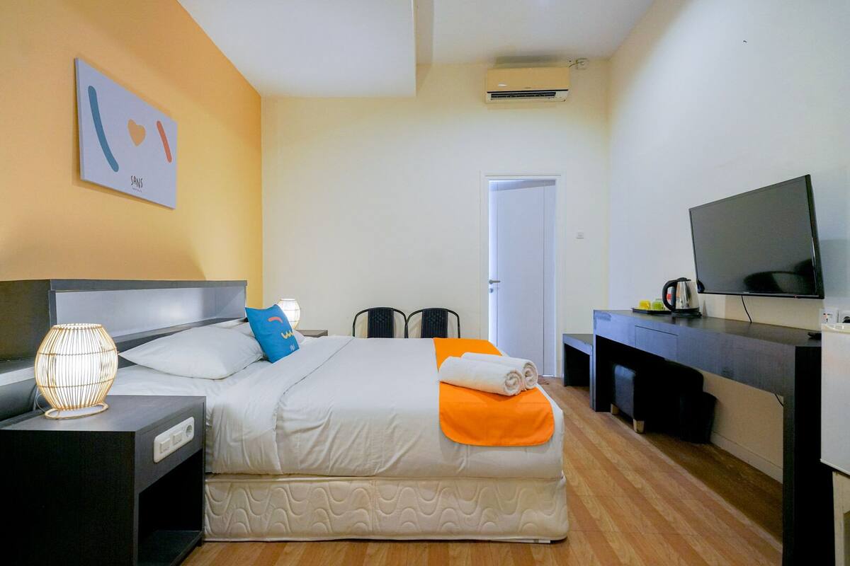 A hotel room with a bed, bedside table, lamp, TV, desk, and an air conditioning unit.