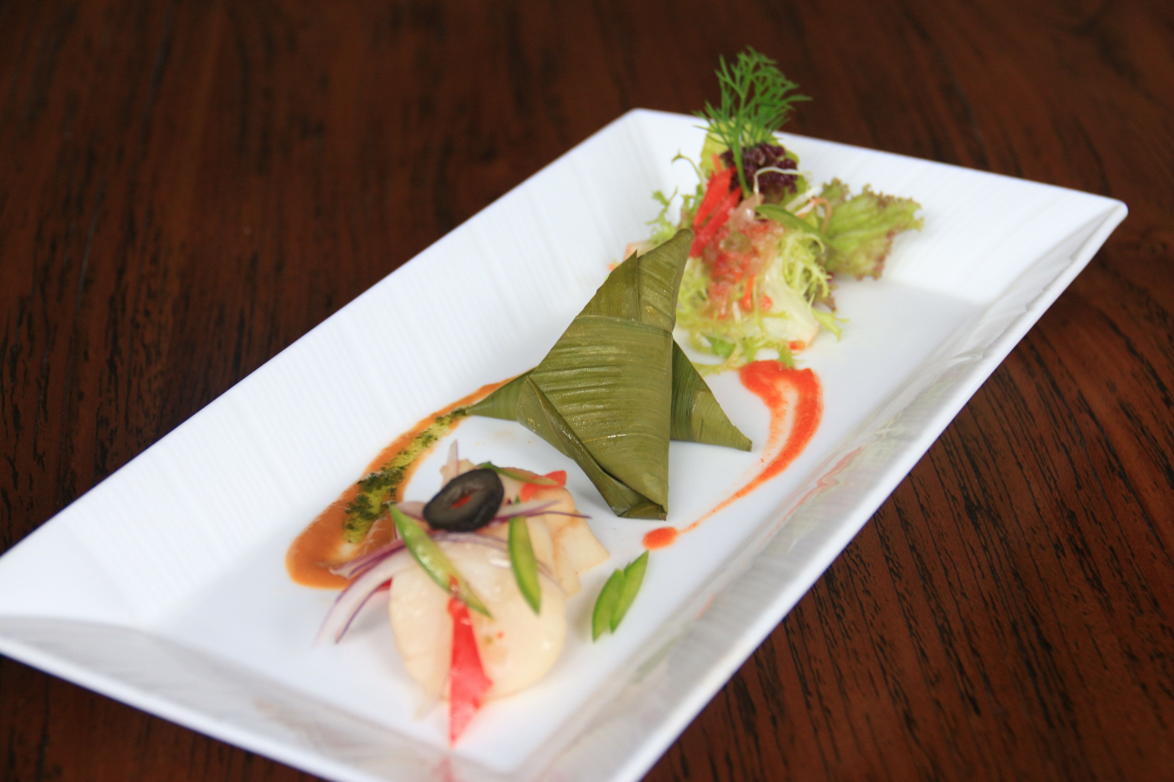 A rectangular white plate with a variety of food items, including a wrapped item, a small salad, and a sauce.