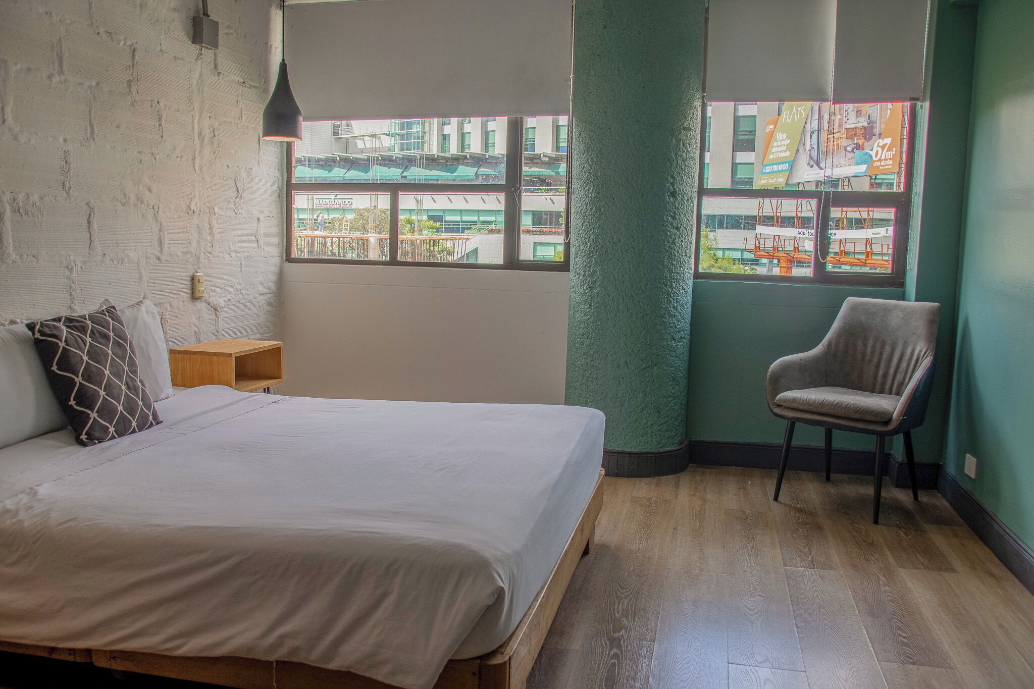 A bedroom with a bed, a chair, a nightstand, a window with a view of buildings, and a textured green wall.