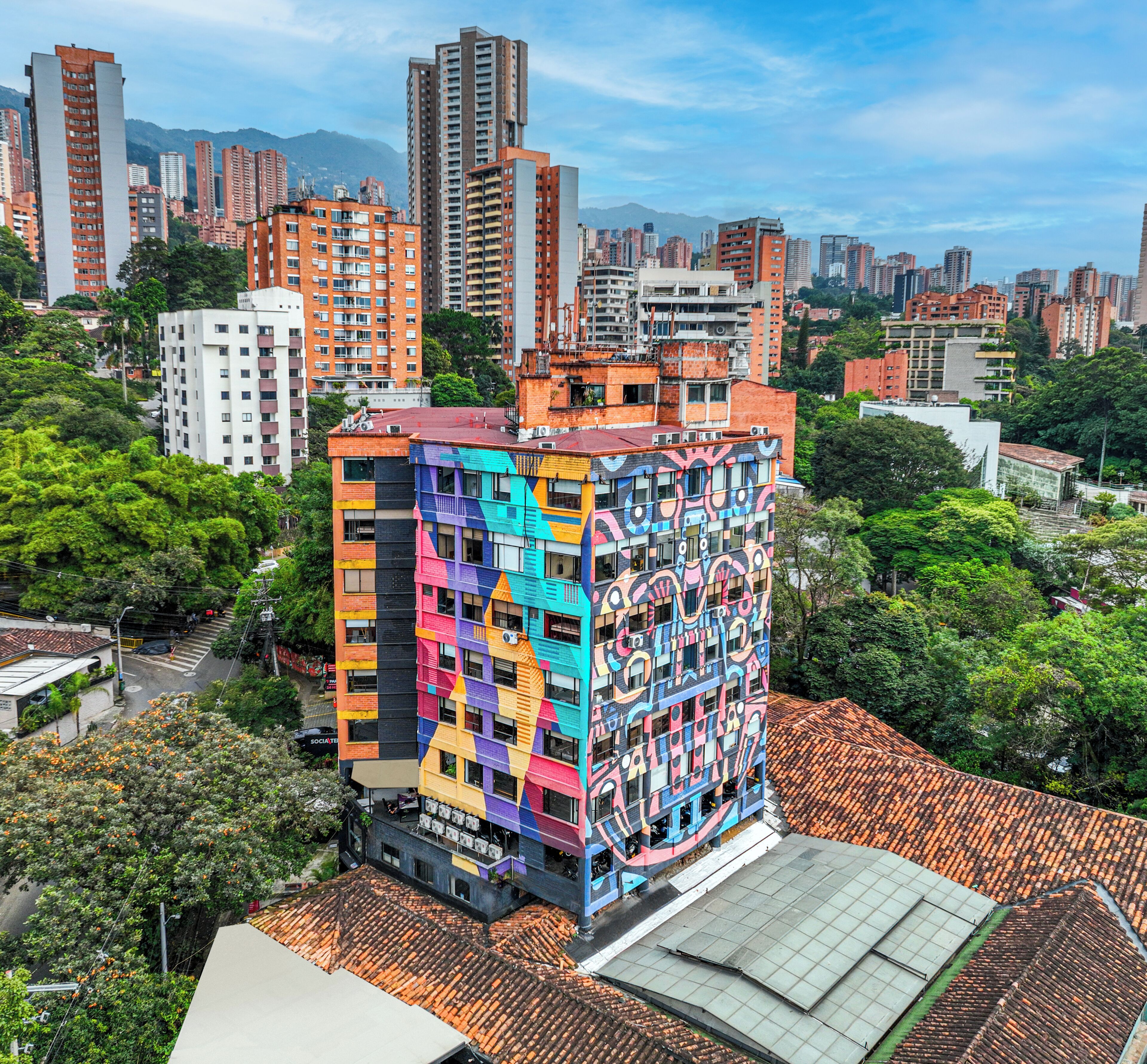 Un edificio de varios pisos con fachada colorida, rodeado de otros edificios altos y vegetación.