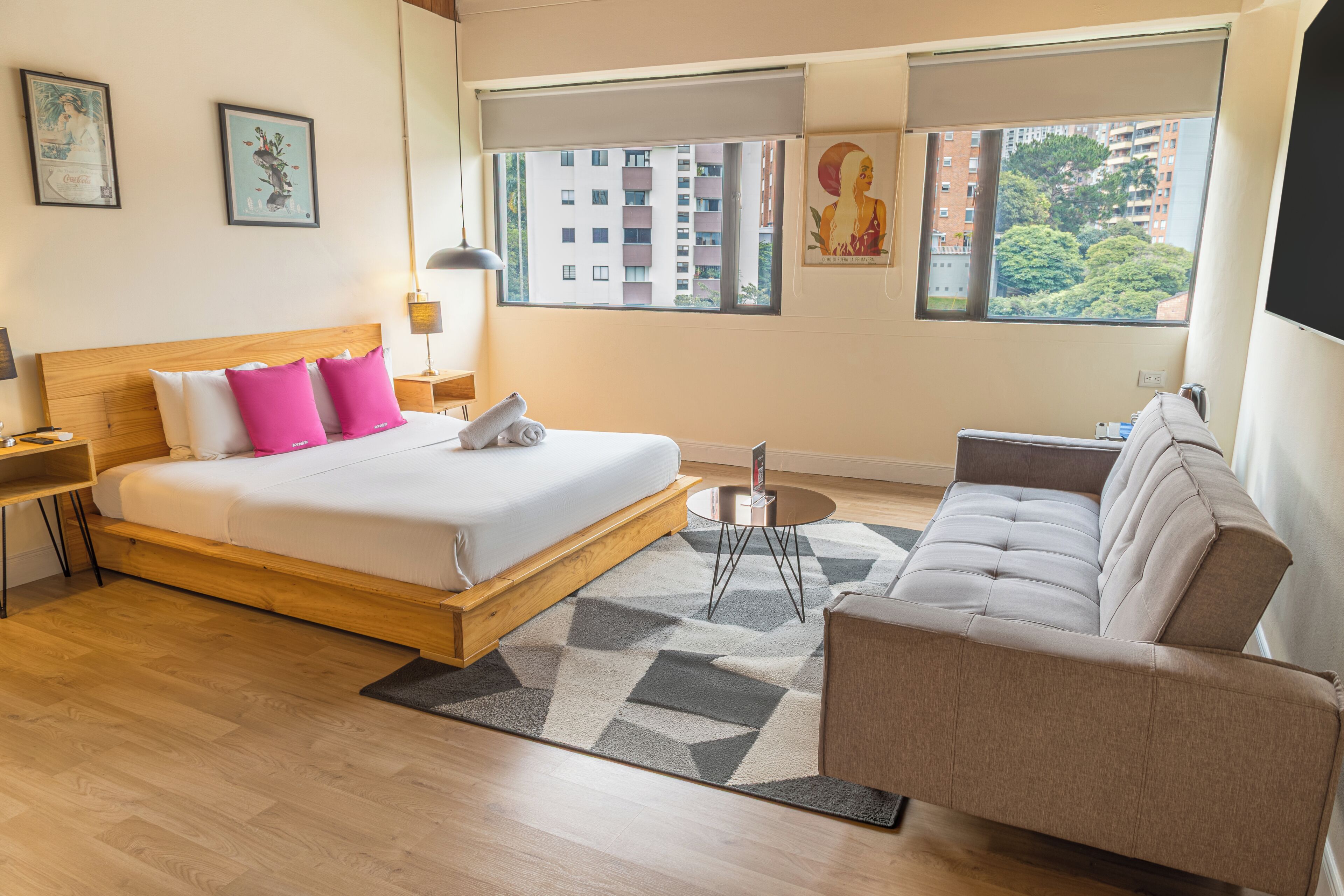 A modern hotel room with a wooden bed, a sofa, a small round table, and a large window offering a city view.