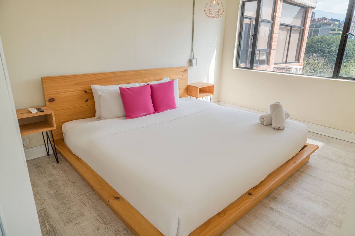 A large bed with a wooden headboard, white bedding, and two pink pillows. There is a bedside table with a lamp, a small wooden stool, and a white towel on the bed.