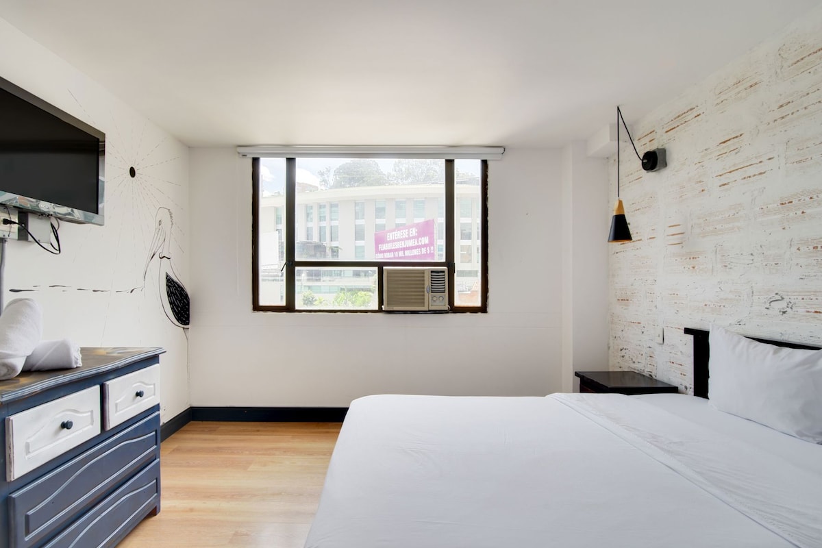 Un dormitorio con una cama grande, un aparador, un televisor montado en la pared y una ventana con vistas a edificios.
