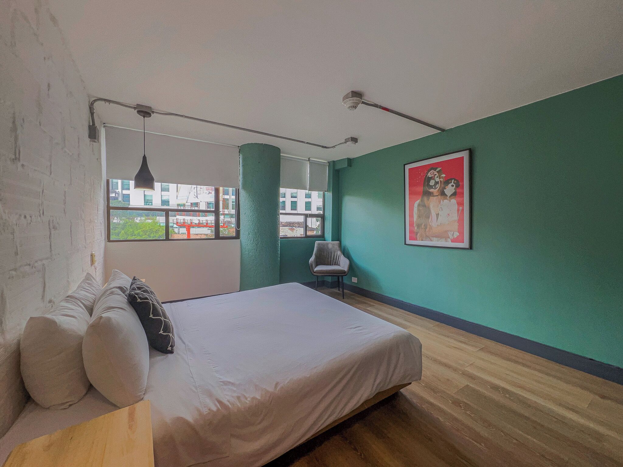 Een slaapkamer met een bed, een stoel, een raam met uitzicht op gebouwen en een schilderij aan de muur.