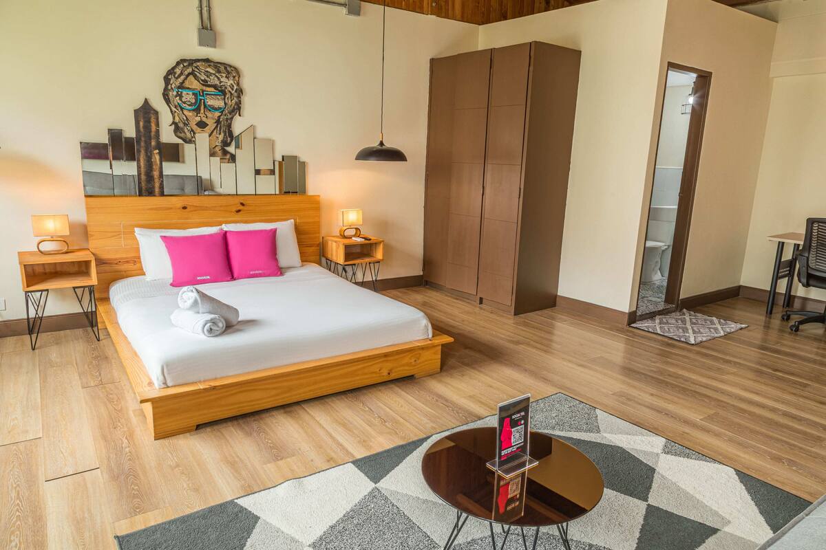 A hotel room with a wooden bed, a headboard with a mirror, a wardrobe, a desk with a chair, and a small table with a lamp.