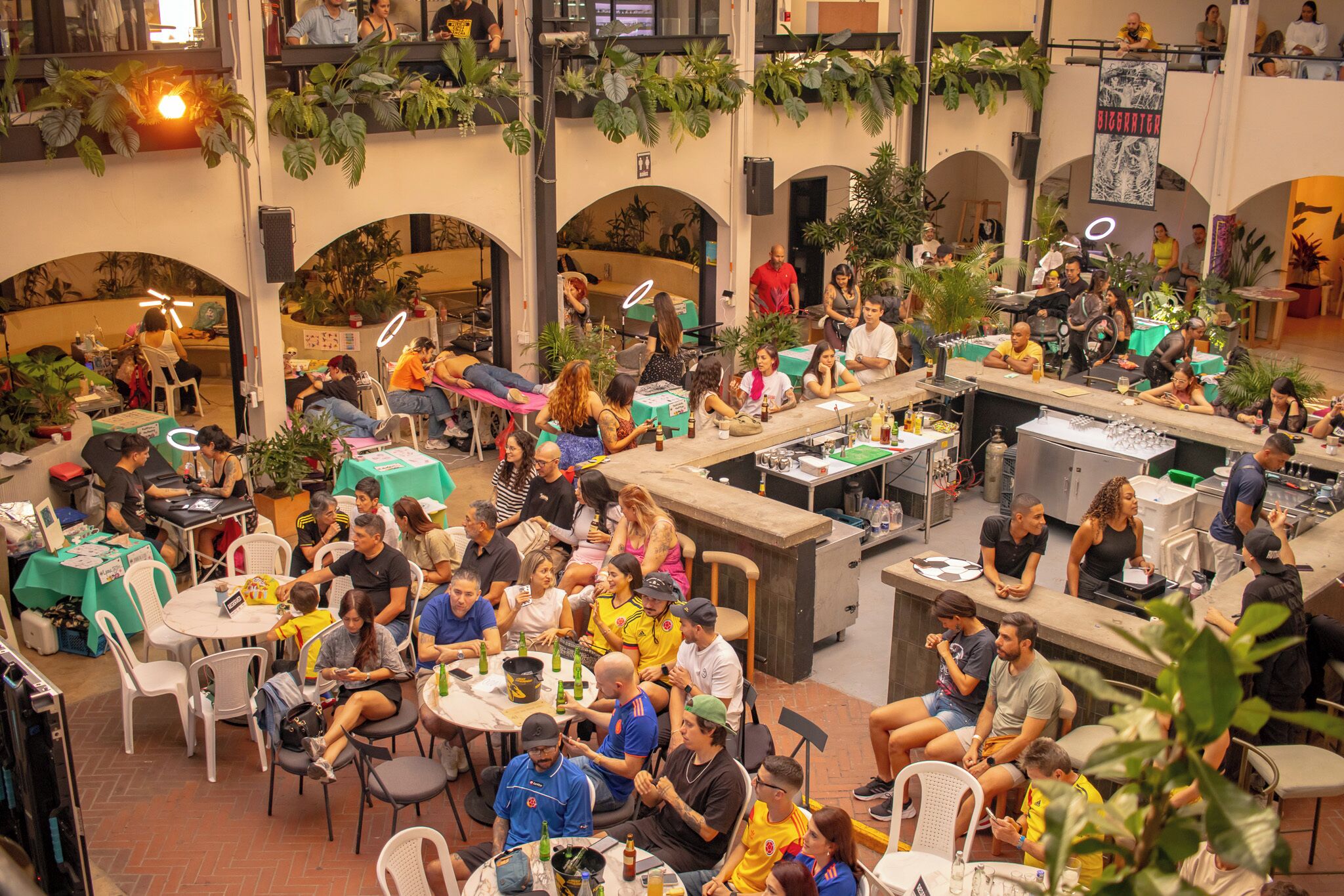 A bustling indoor social space with multiple tables and seating areas, surrounded by greenery and architectural arches.