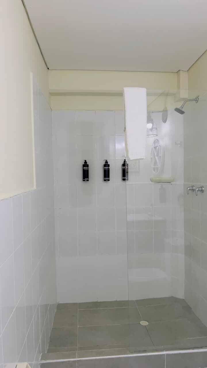 Cabina doccia con pareti in vetro, un asciugamano bianco appeso al muro, soffione doccia in vetro e due flaconi di shampoo a parete.