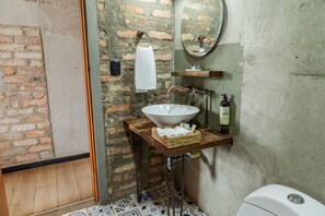 A bathroom with a rustic brick wall, a white sink on a wooden shelf, a towel hanging on a towel ring, a mirror, and a toilet. - Delgadillo Boutique Hotel by Socialtel (Bogotá)