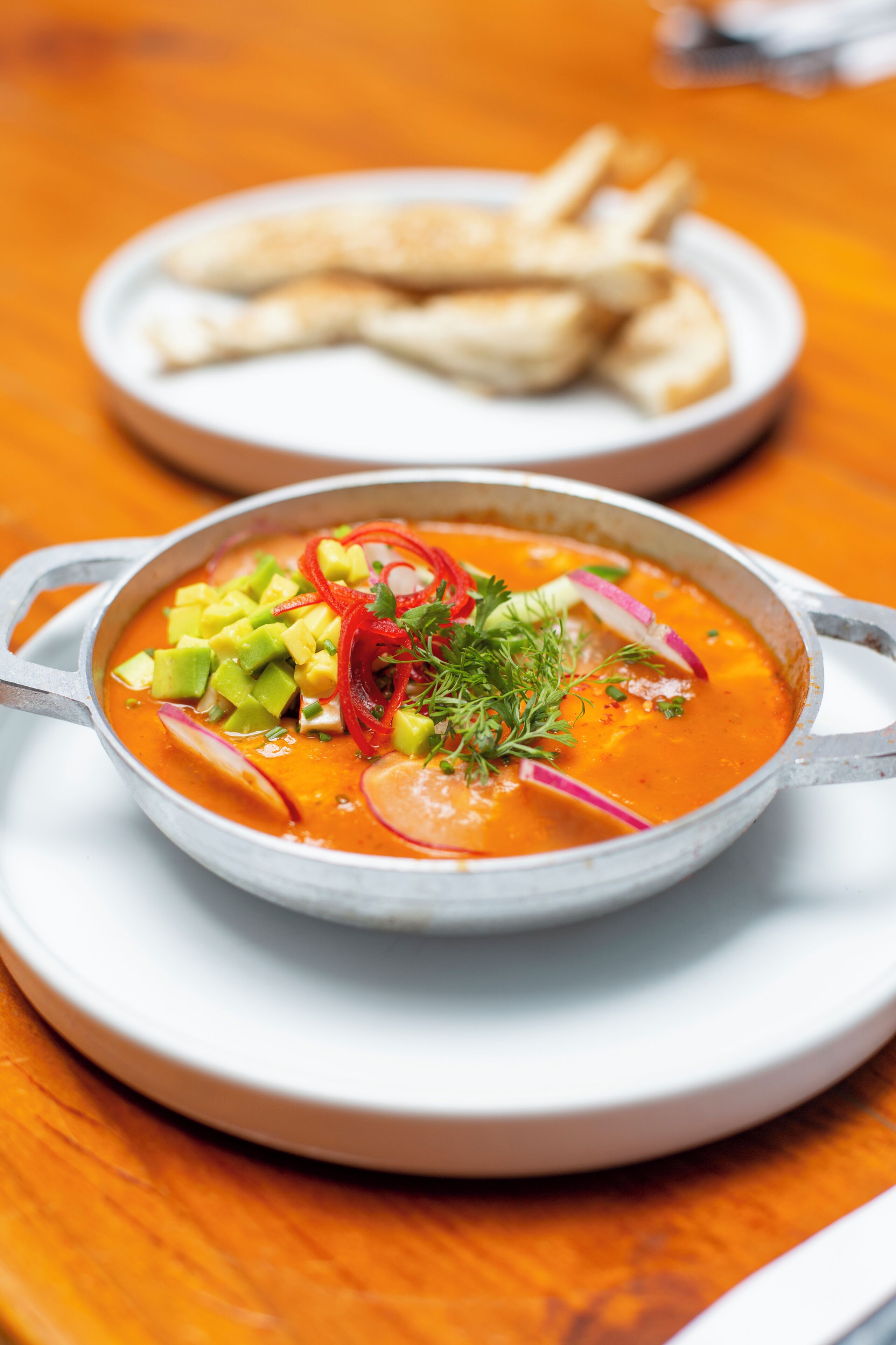 A bowl of soup with avocado and red peppers on a white plate.