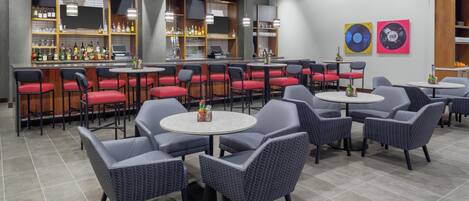 Zona de bar moderna con mostrador, taburetes y mesas redondas con sillas.