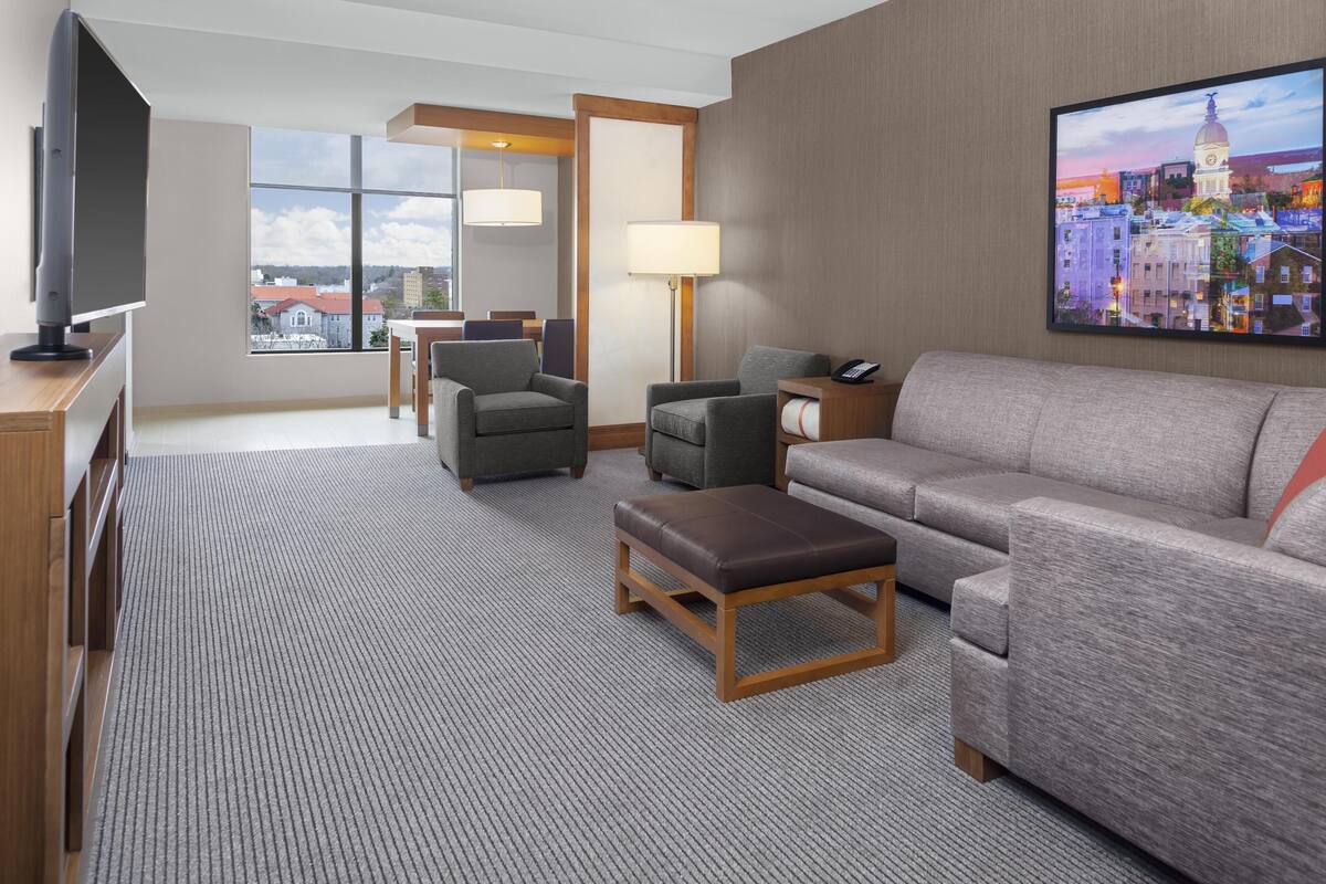 Habitación de hotel con un ventanal grande, un televisor de pantalla plana, un sofá, dos sillones, una mesa de centro y una lámpara.