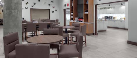A modern cafe with round tables and chairs, a wooden bookshelf, and a kitchen area visible through an open door.