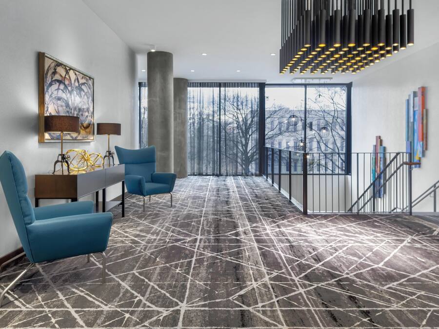 A modern hotel lobby with a geometric patterned floor, blue seating, a large abstract painting, and a staircase with a railing.