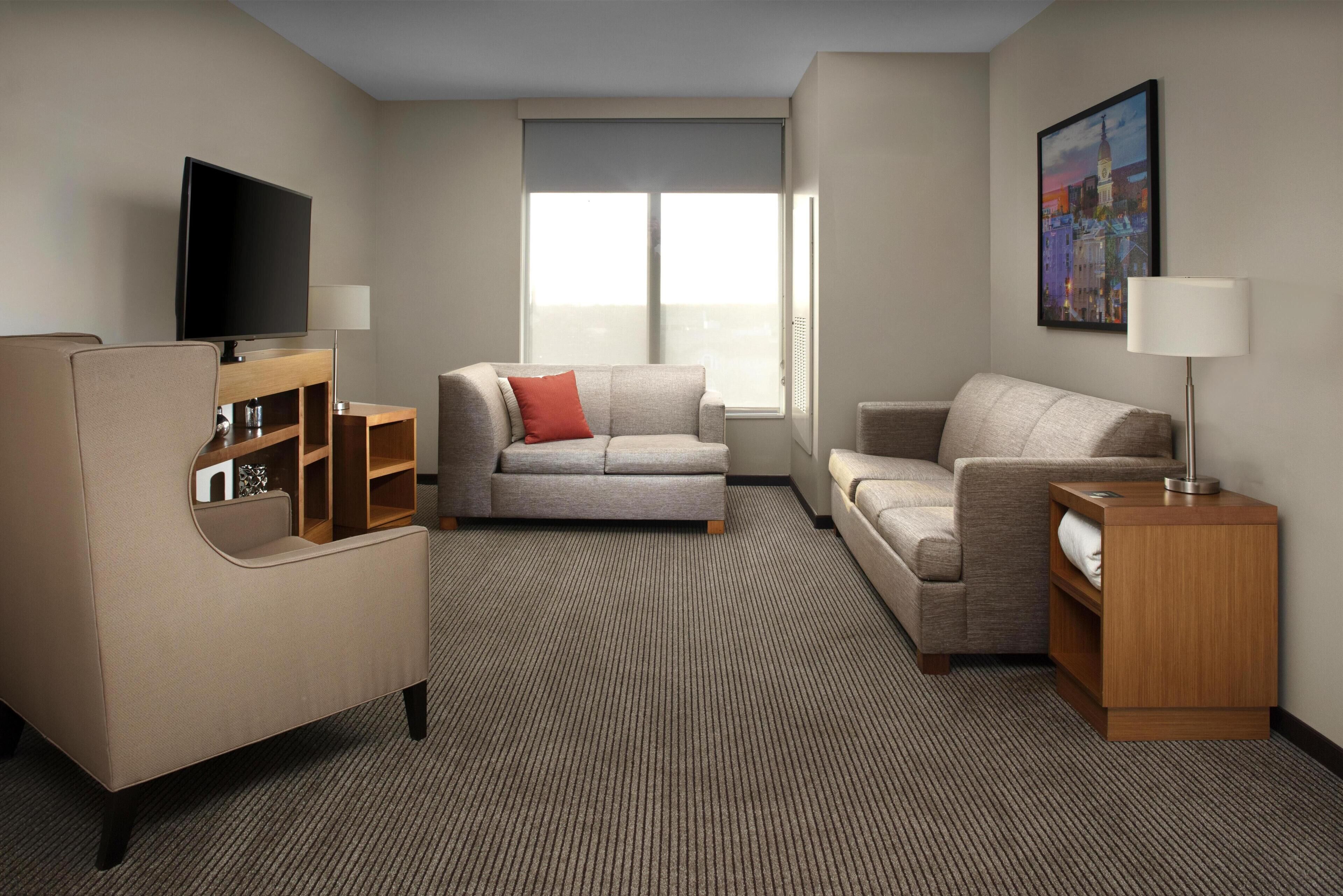 A hotel room with a flat-screen TV, two sofas, a wooden side table, and a painting on the wall.
