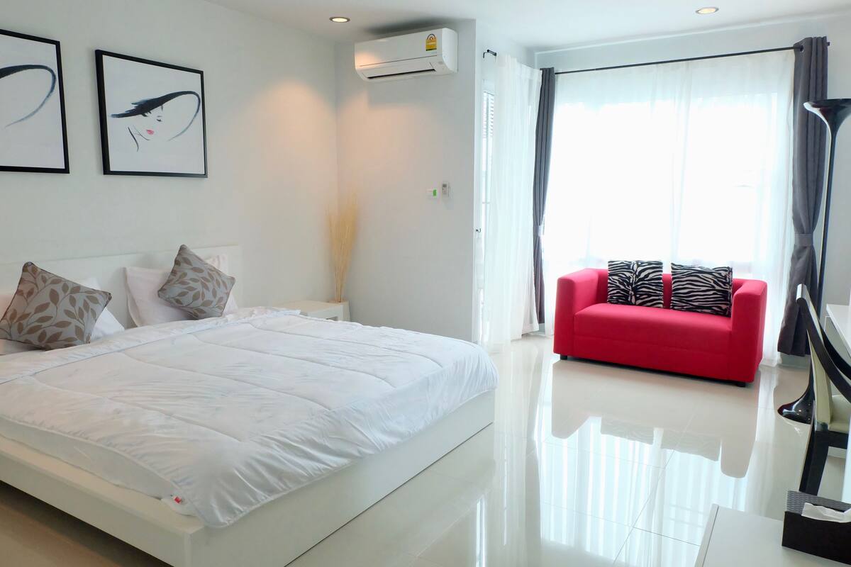 A modern hotel room with a large bed, a red sofa, and a large window with curtains.