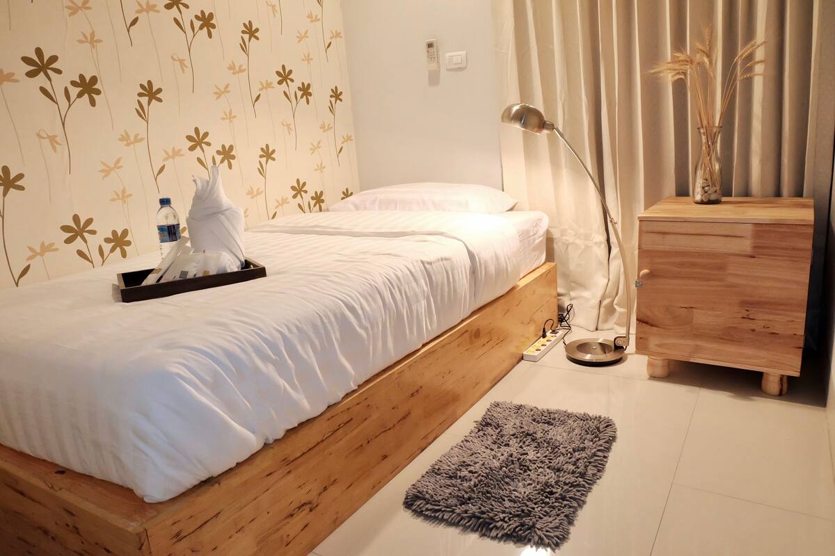 A bedroom with a wooden bed, a bedside table with a lamp, a small wooden dresser, and a grey rug.
