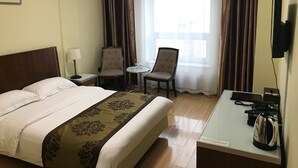 Ett hotellrum med en säng, ett skrivbord med en vattenkokare, en stol, ett litet bord med en lampa och ett fönster med gardiner.