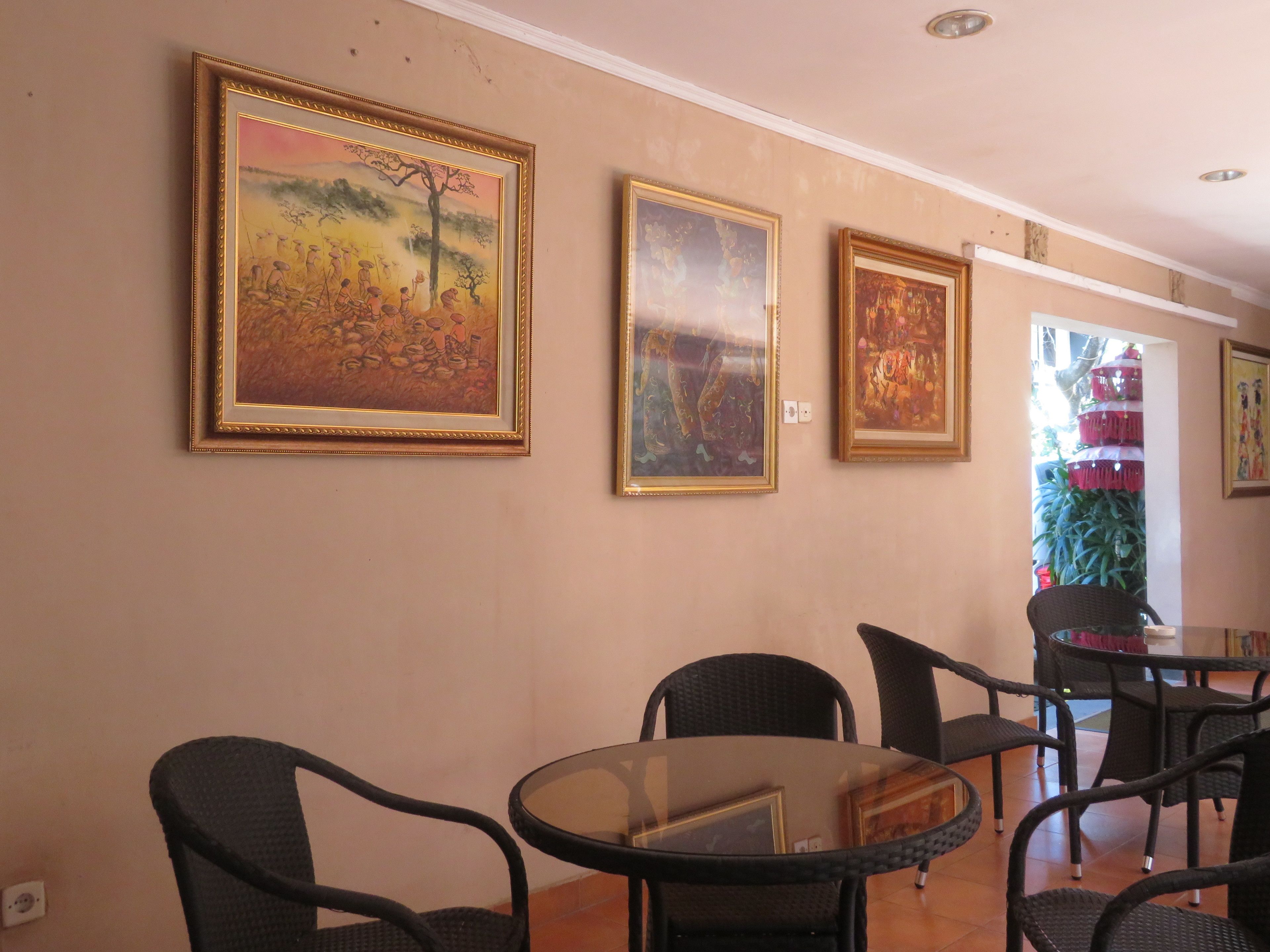 A dining area with wicker chairs and a glass table, framed paintings on the wall, and a view of a plant outside.