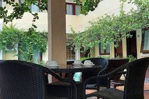 An outdoor seating area with wicker furniture and tables, surrounded by greenery and a building facade. - Balira Airport Hotel (Tuban)