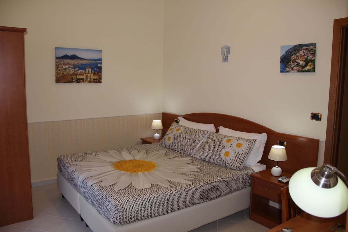 Una cama bien hecha con un edredón floral grande, dos mesitas de noche con lámparas, y dos cuadros en la pared que muestran paisajes.