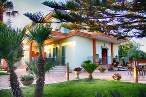 Uma casa de dois andares com varanda, rodeada por palmeiras e plantas em vasos.