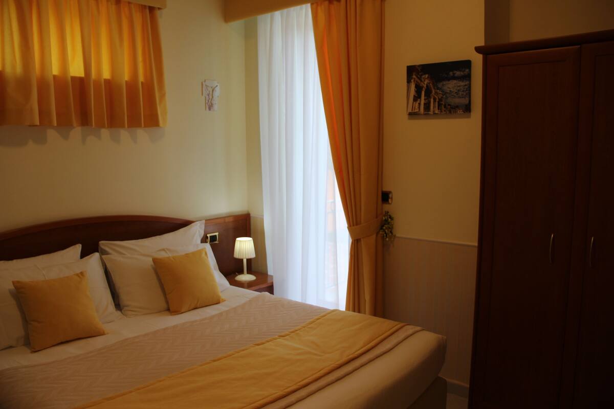 Habitación de hotel con una cama grande, una mesita de noche con lámpara, un armario y una ventana con cortinas.