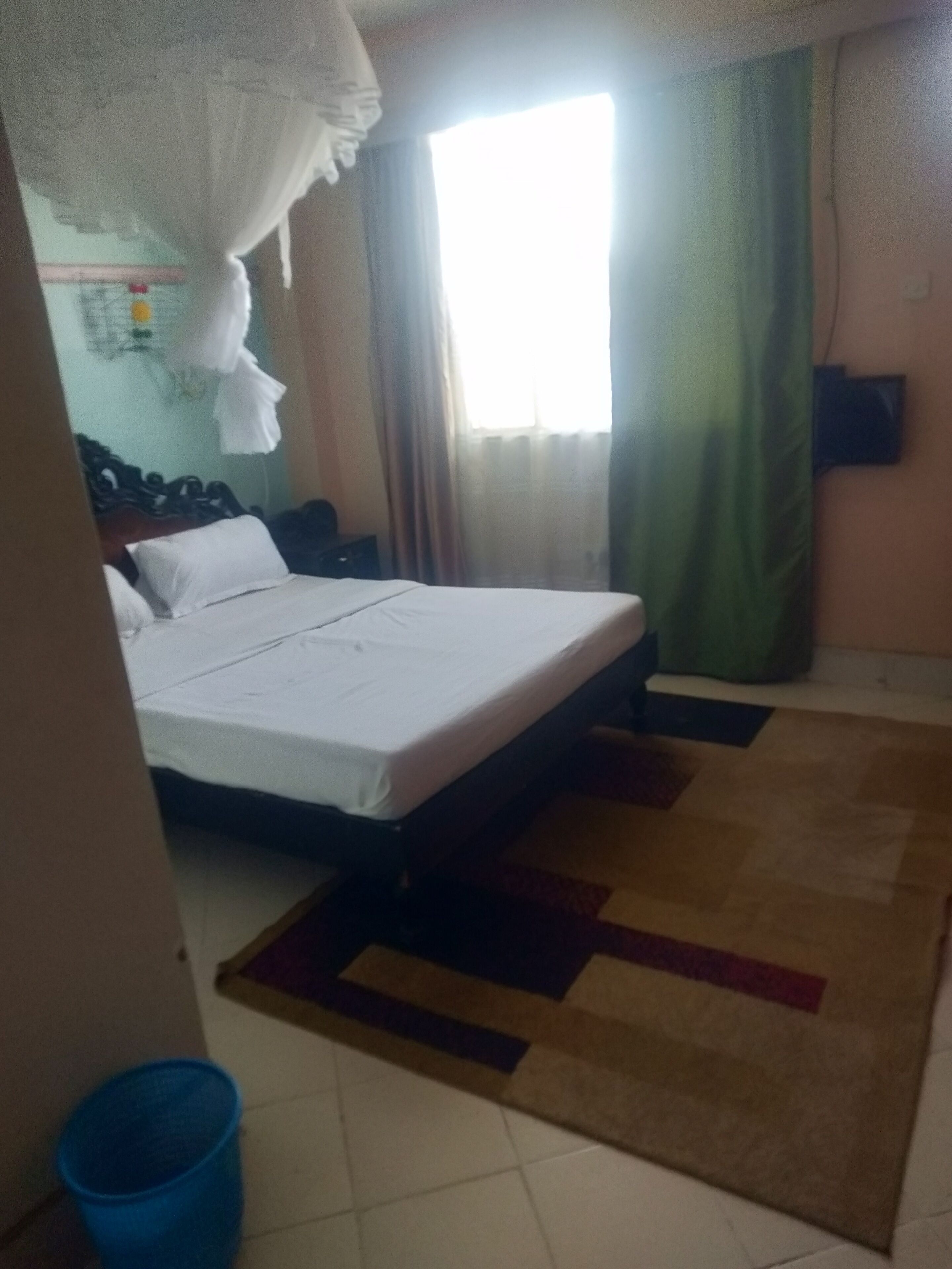 Una habitación de hotel con una cama, una mesita de noche, un cubo de basura azul y una ventana con cortinas.
