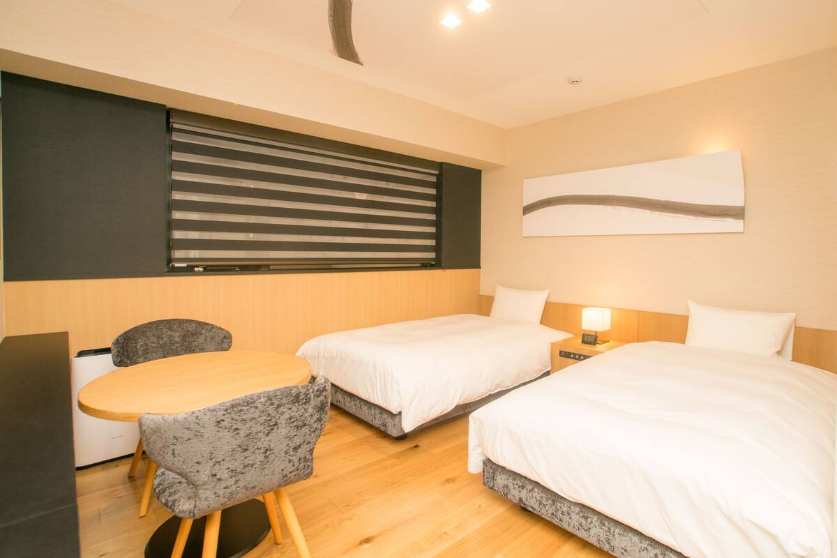 A modern hotel room with a wooden floor, a desk with a chair, two beds, a large window with blinds, and a wall-mounted abstract art piece.