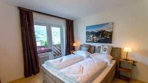 A double bed with white linens, a nightstand with a lamp, a painting of a town on the wall, and a window with curtains. - SKI-N-LAKE City Apartments (Zell am See)