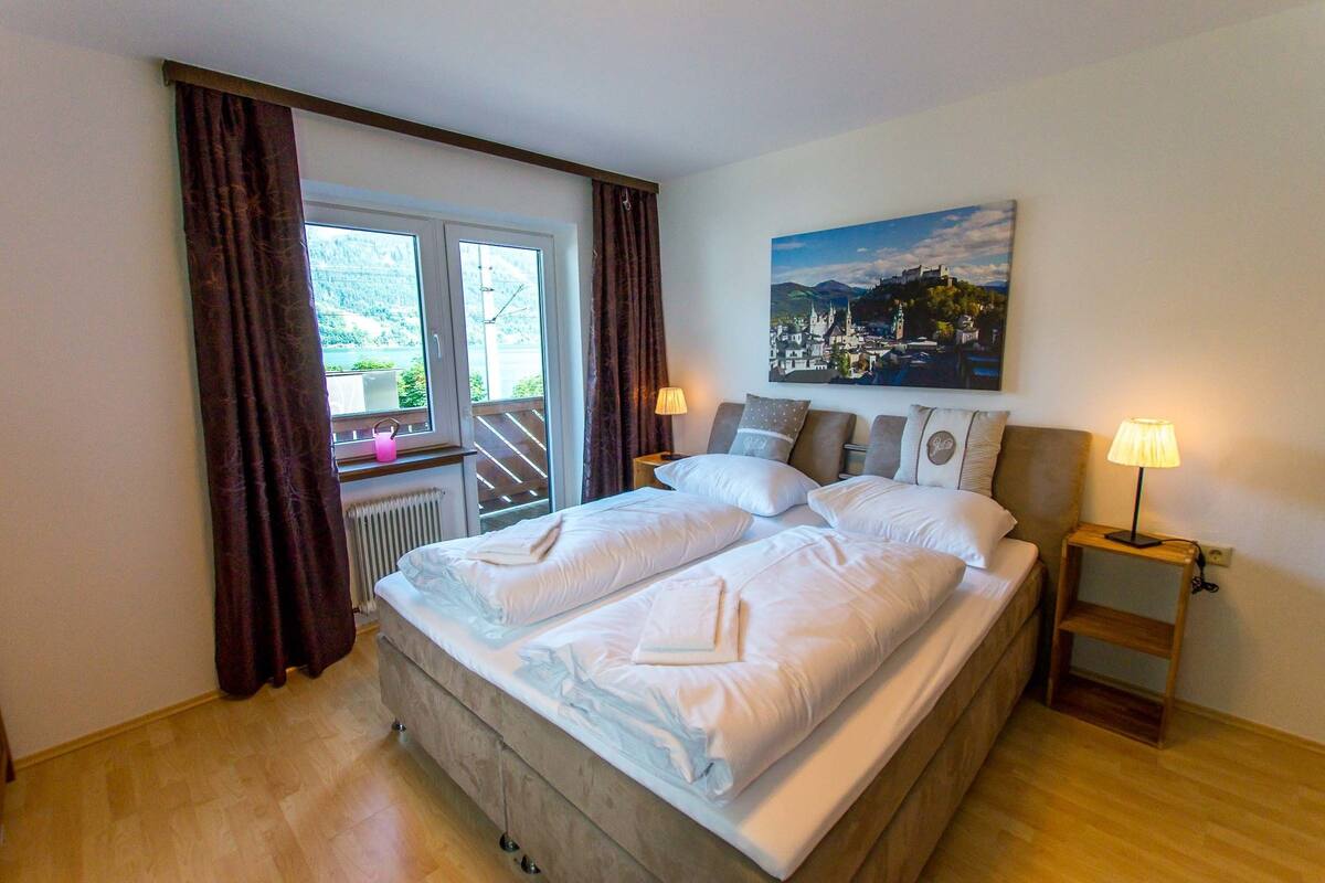 A double bed with white linens, a nightstand with a lamp, a painting of a town on the wall, and a window with curtains.