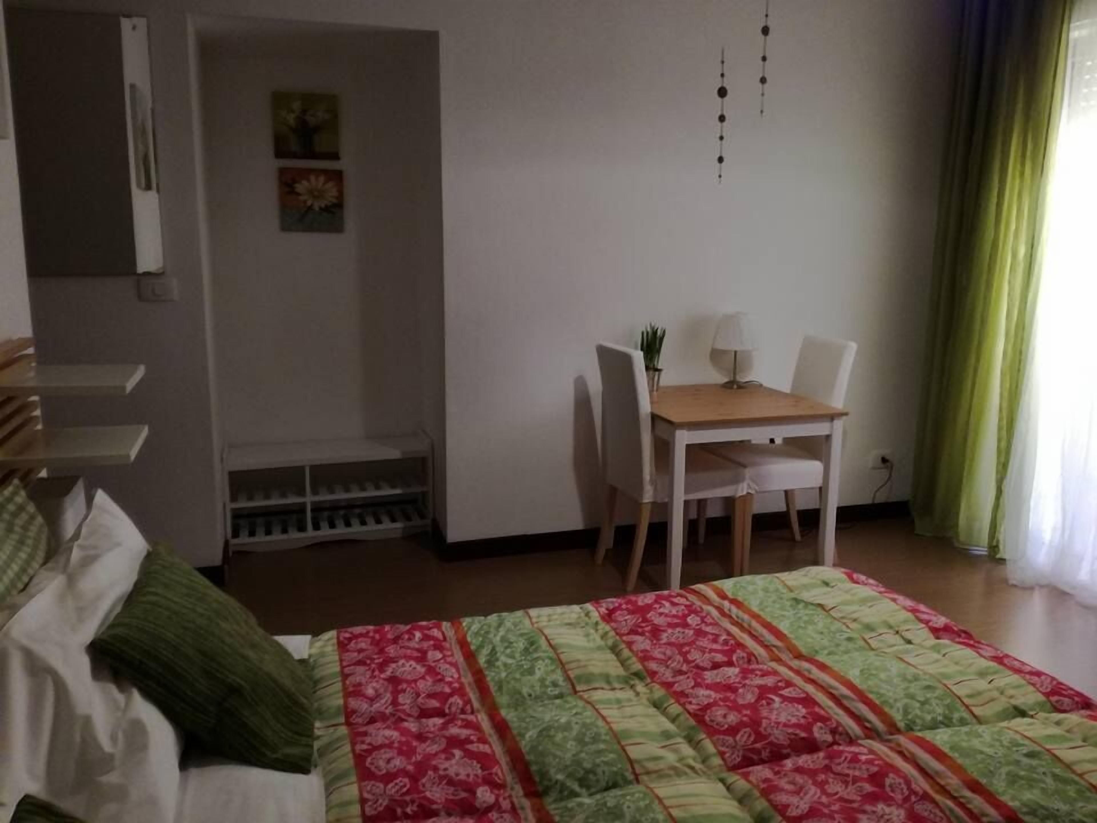 A room with a bed, a desk, and a chair. There is a window with curtains, a wall with a picture, and a shelf with items.