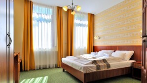 Una habitación de hotel con una cama de madera, una alfombra verde estampada, un armario de madera y una ventana con cortinas amarillas.