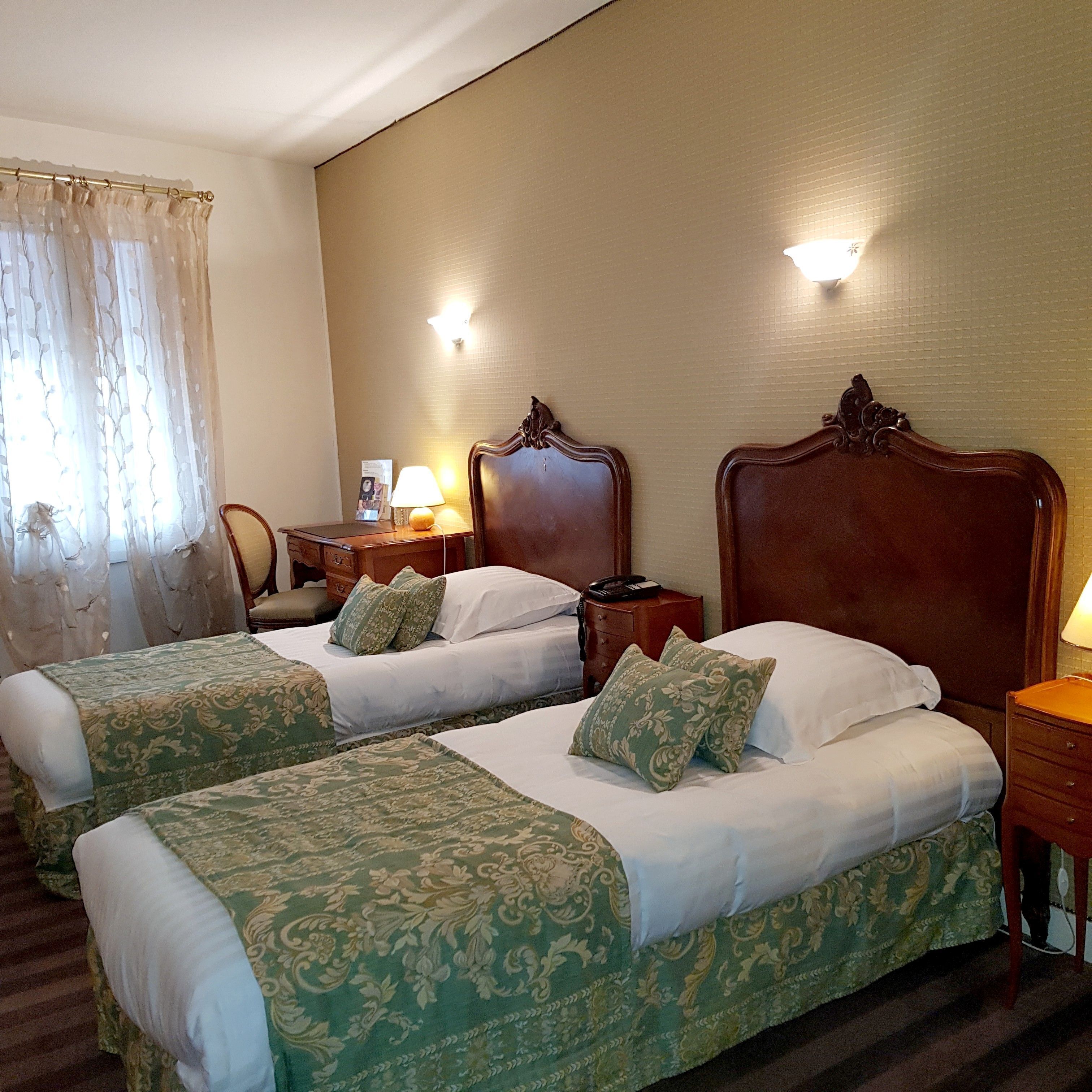 A hotel room with two beds, each with floral-patterned bedding, a wooden headboard, bedside tables with lamps, and a window with sheer curtains.