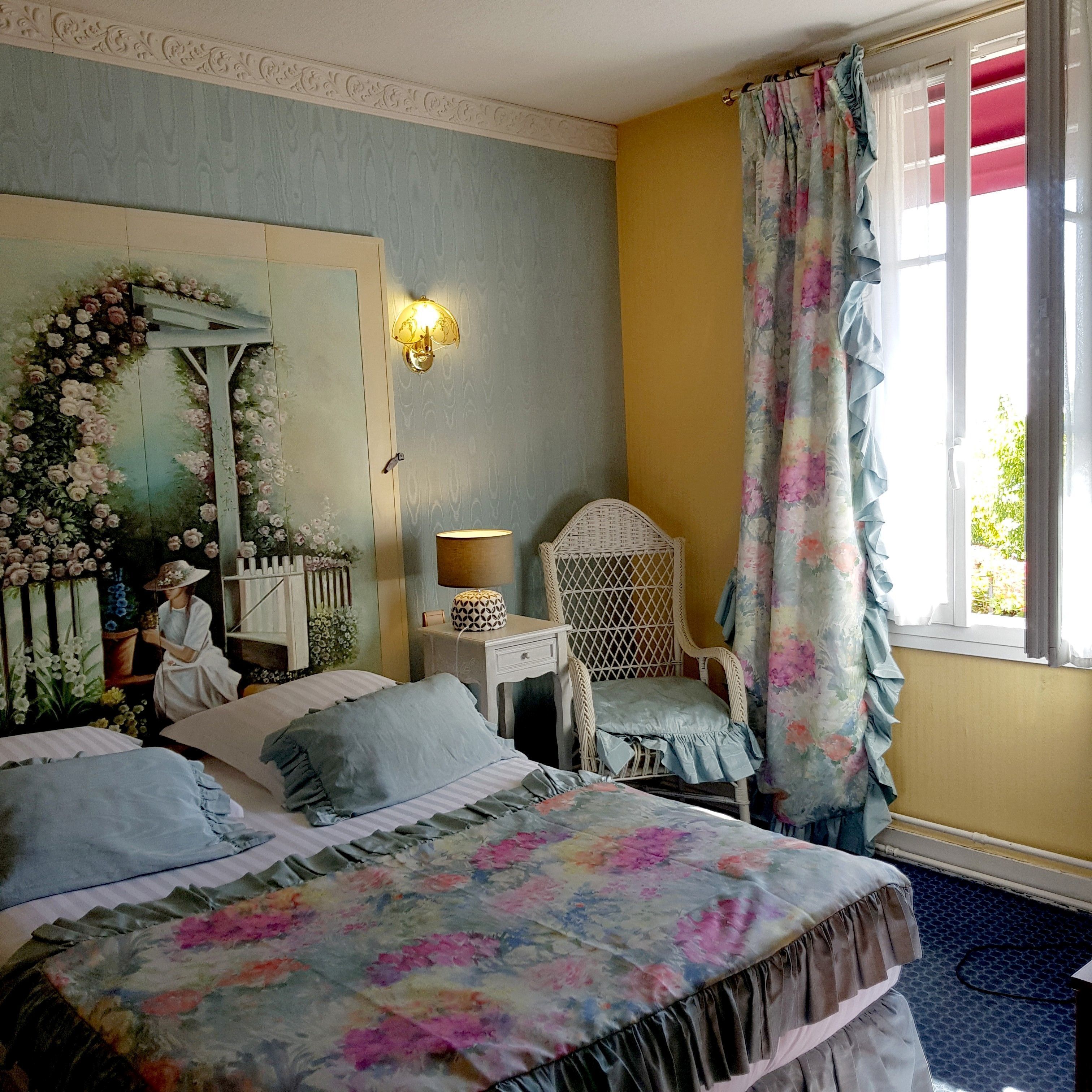 Une chambre à coucher avec une couvre-lit à motifs floraux, une chaise blanche, une table de chevet avec une lampe et un miroir décoratif orné de motifs floraux.