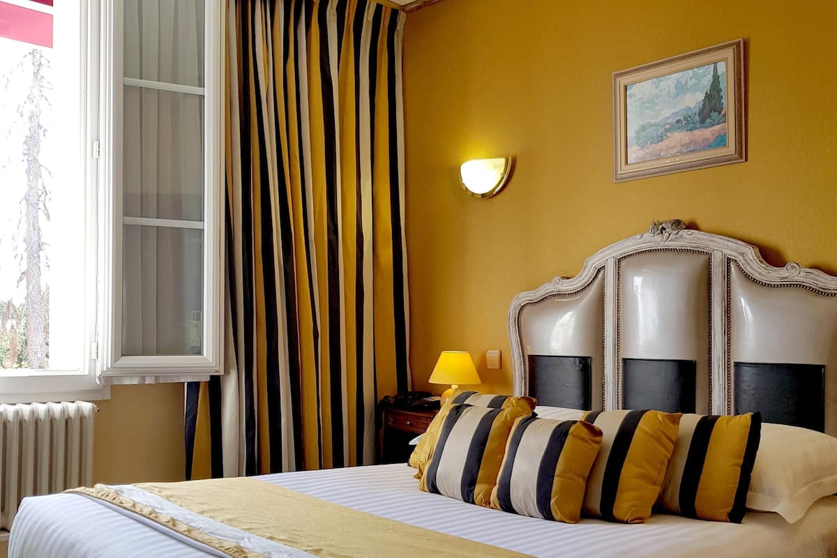 A hotel room with a large bed, a nightstand, a lamp, a painting on the wall, and a window with curtains.