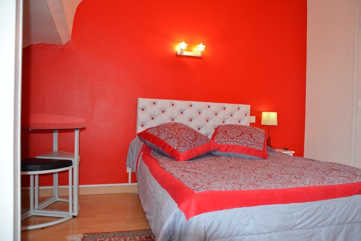 A bedroom with a red wall, a tufted white headboard, two bedside lamps, and a small round table with a chair.