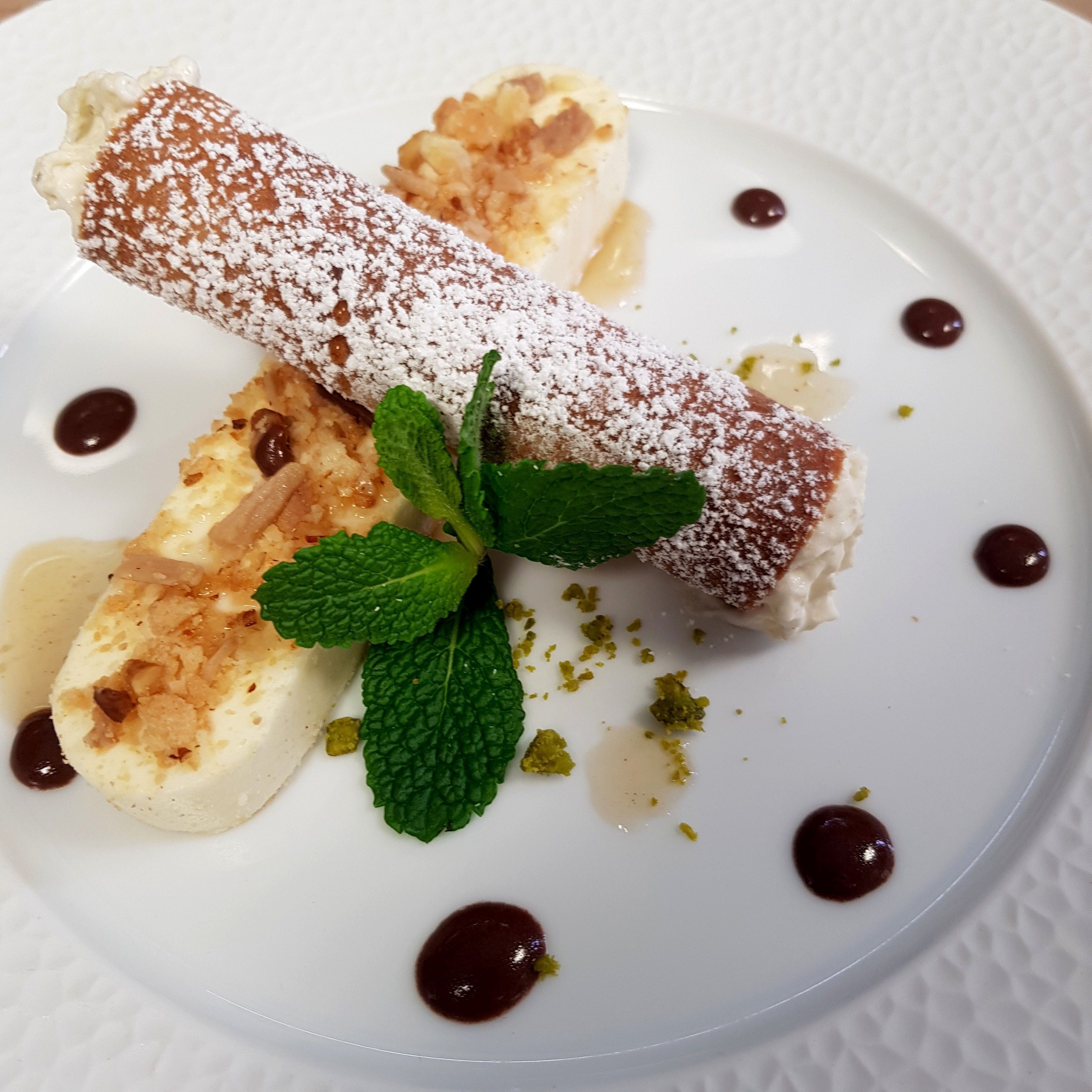 Een nagerechtenbord met een gerold gebakje bestrooid met poedersuiker, vergezeld van een muntblaadje en chocoladesaus.