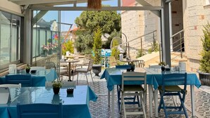 An outdoor dining area with tables and chairs, covered by a white roof, and overlooking a courtyard with trees and buildings. - Allegria Hotel (Cesme)