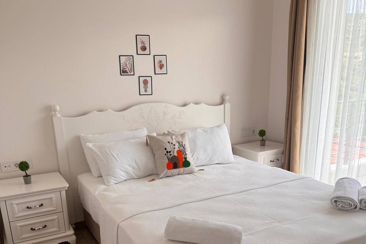 A neatly made bed with white linens and a rolled towel, a white nightstand with a small plant, and a wall with framed artwork.
