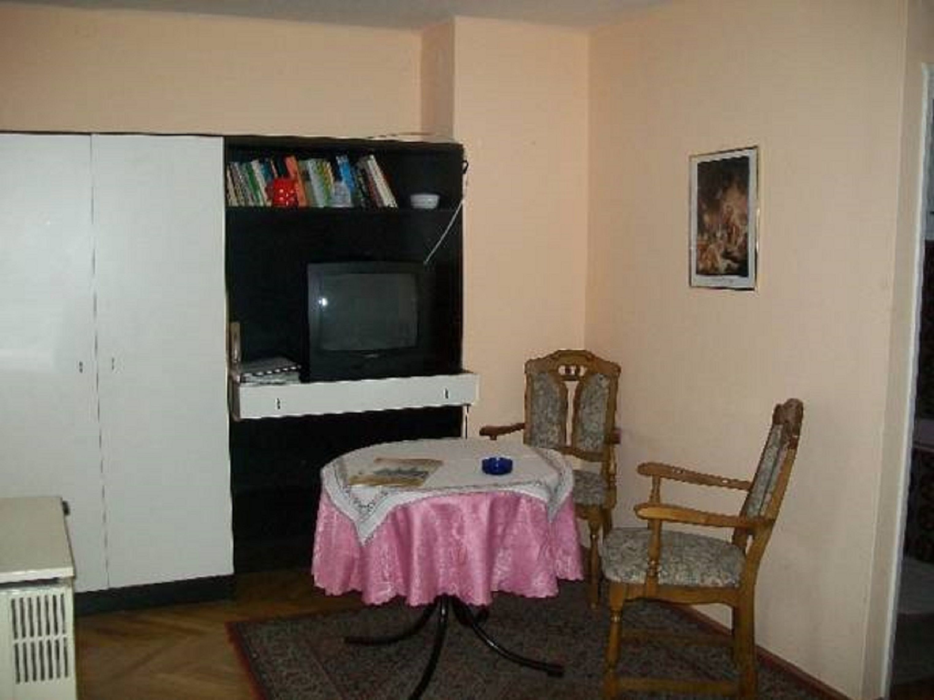 Una habitación pequeña con una televisión en un estante, una mesa con mantel rosa y dos sillas.