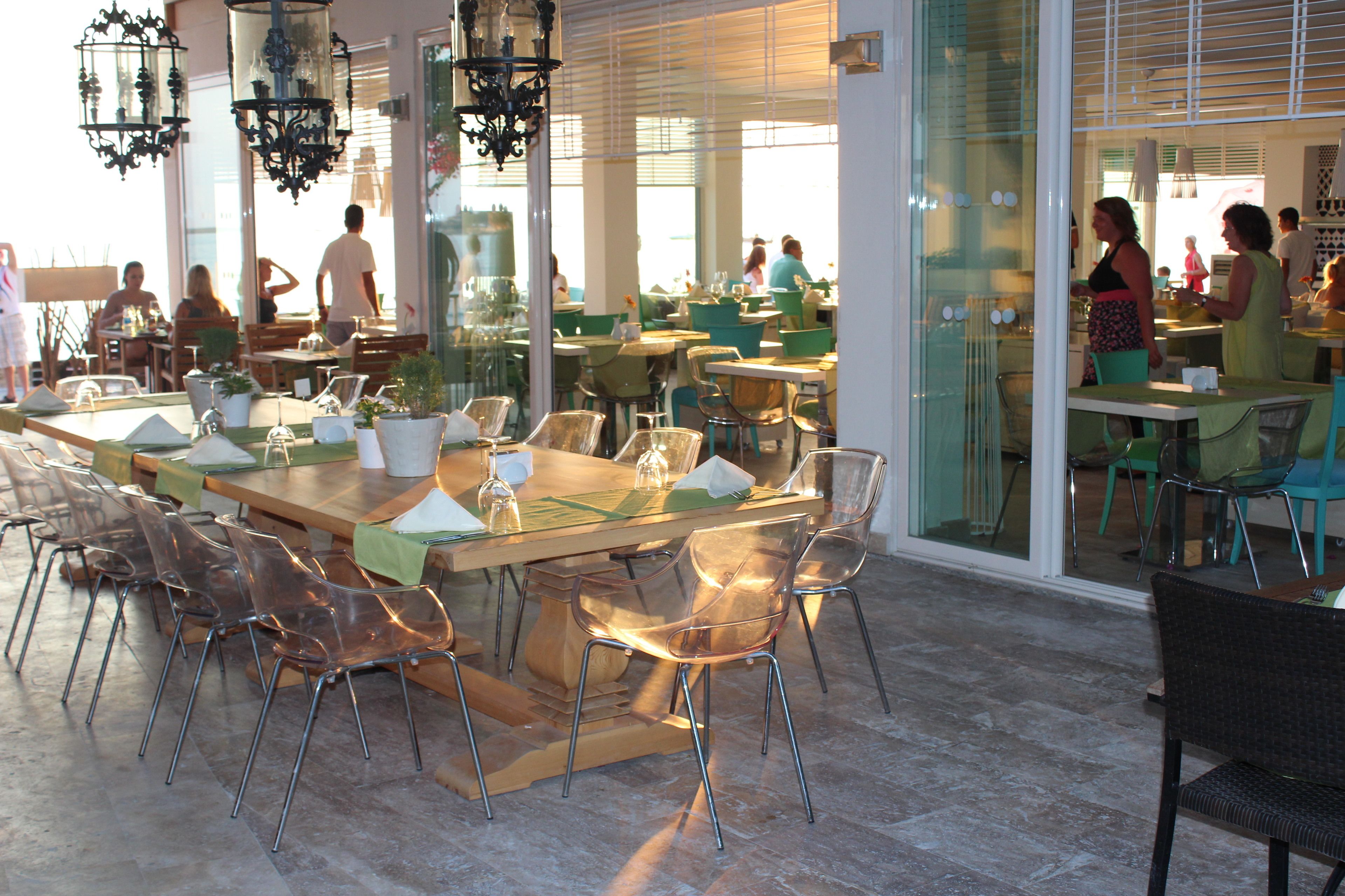 Een ruime restaurantruimte met meerdere eettafels en stoelen, grote ramen en hangende lampen.