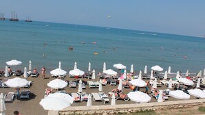 A beach with numerous white umbrellas and sun loungers, a clear blue sky, and a calm sea with boats and swimmers.
