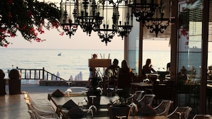 A restaurant with a view of the sea, featuring outdoor seating with clear chairs and tables, potted plants, and hanging light fixtures.