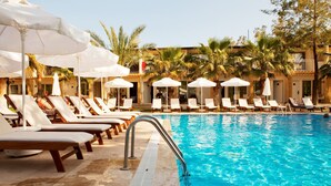 A poolside area with lounge chairs and umbrellas, surrounded by palm trees and a building with balconies. - Sunprime Dogan Side Beach - Adults Only (Manavgat)