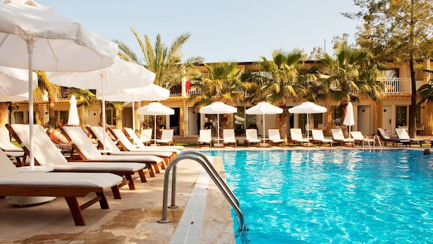 A poolside area with lounge chairs and umbrellas, surrounded by palm trees and a building with balconies.