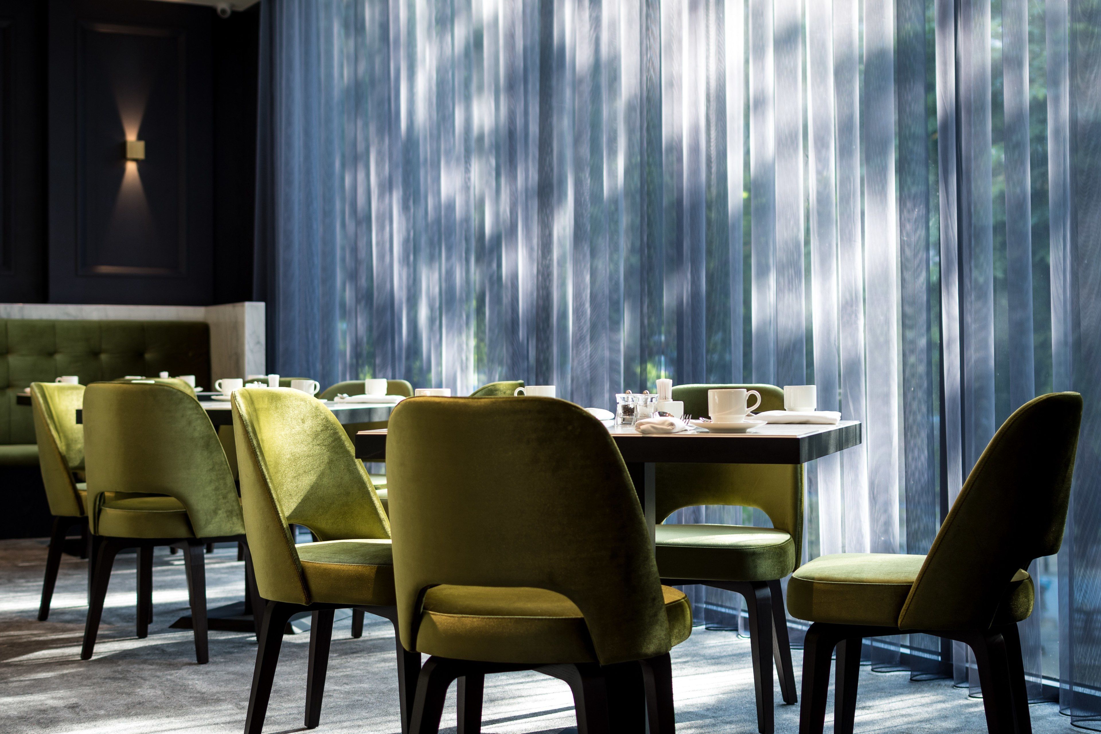 A dining area with green upholstered chairs and tables, a tufted green sofa, and a view through sheer curtains.