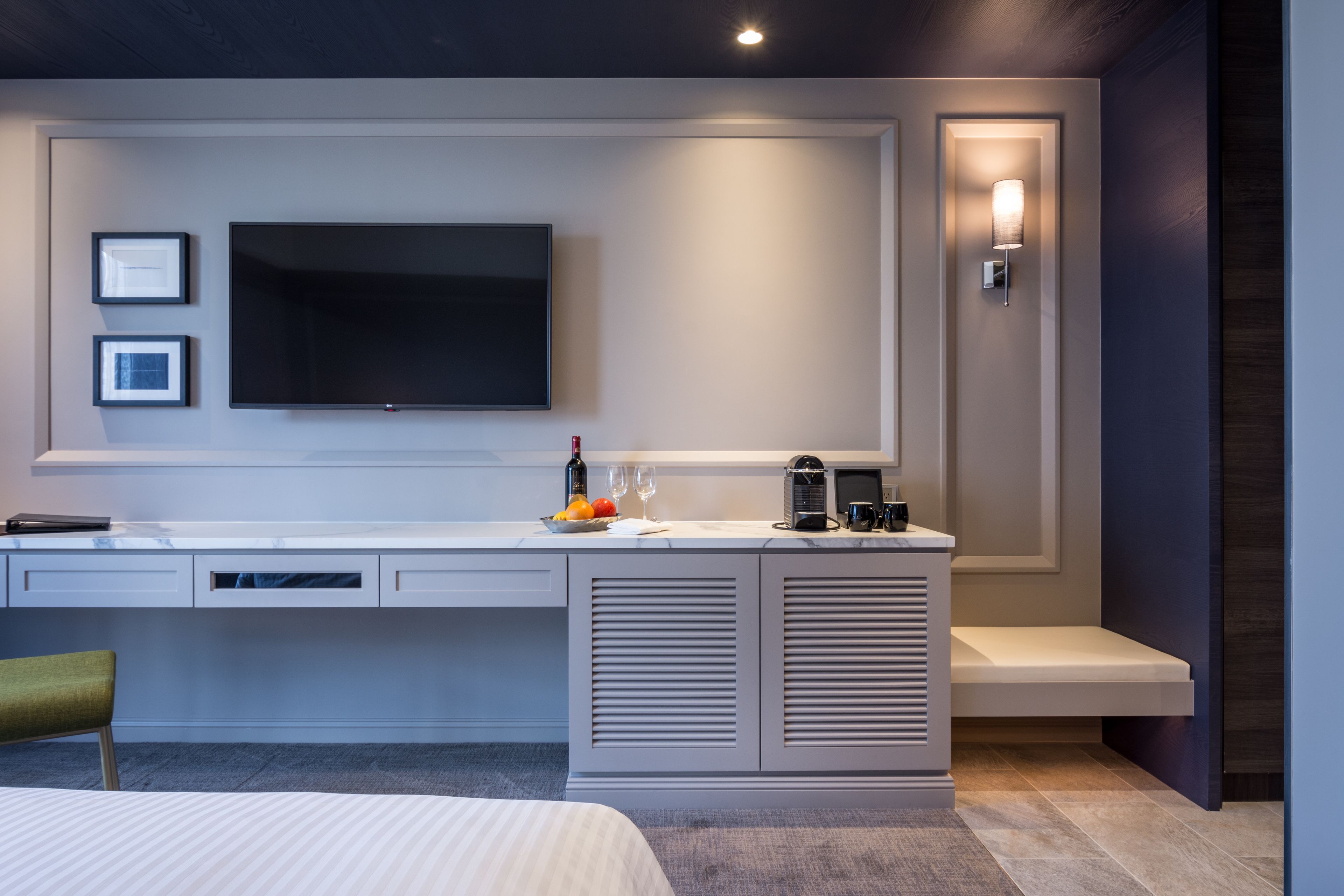 A modern hotel room with a large flat-screen TV mounted on the wall, a desk with a laptop, a coffee maker, and a small seating area.