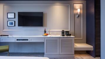 A modern hotel room with a large flat-screen TV mounted on the wall, a desk with a laptop, a coffee maker, and a small seating area.